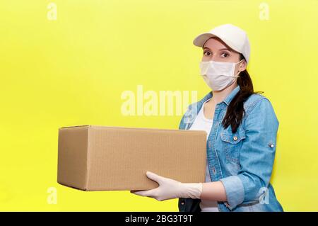 Livraison en toute sécurité. Femme Courier dans masque de protection et gants avec boîte en carton sur fond jaune. Concept anti-coronavirus. Banque D'Images