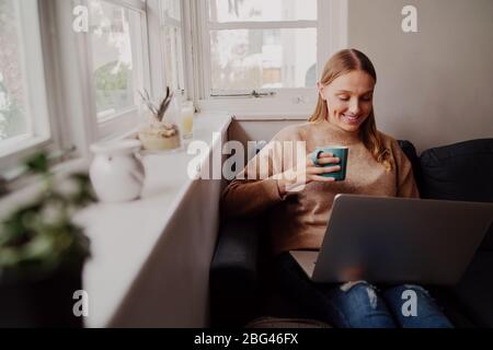 Jeune femme souriante sous l'isolation de soi en profitant du café du matin en utilisant un ordinateur portable assis à la maison sur un canapé Banque D'Images