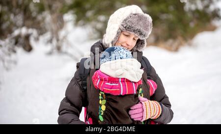 Une jeune mère heureuse transportant son bébé dans un porteur lorsqu'ils traversent une terre d'hiver. Banque D'Images