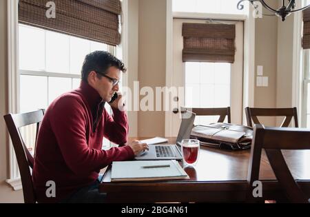 Homme sur un téléphone portable travaillant à la maison en utilisant un ordinateur à une table à manger. Banque D'Images