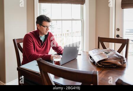 Homme dans des lunettes travaillant à la maison en utilisant un ordinateur à une table à manger. Banque D'Images