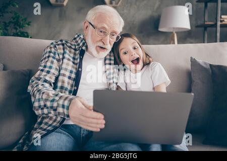 Photo de grand-père d'âge fou jolie petite petite-fille asseyez canapé séjour maison quarantaine regarder comédie humoristique carnet de film bonne humeur moderne design Banque D'Images