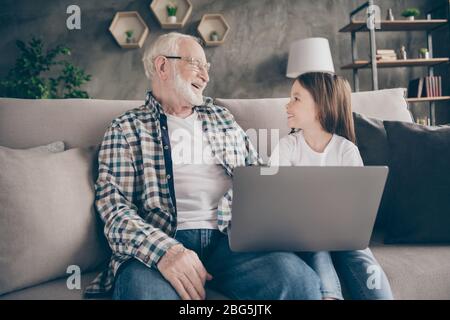 Photo de drôle vieux grand-père petite-fille assis canapé séjour maison quarantaine sécurité regarder comédie humoristique film carnet parler skype moderne Banque D'Images