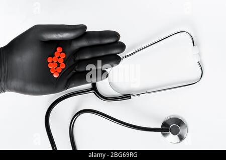 Stéthoscope et gant médical noir contenant des pilules rouges sur fond blanc. Frais généraux Banque D'Images