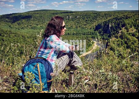 Jeune femme heureuse avec un sac à dos se trouve sur une falaise et regarde la vallée en dessous Banque D'Images