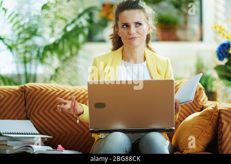 triste femme d'apprenant moderne en jean et veste jaune dans la maison moderne en journée ensoleillée étude en ligne sur un ordinateur portable. Banque D'Images