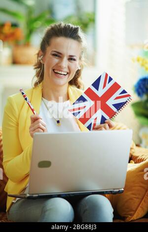 Femme souriante de 40 ans, en Jean et en veste jaune, avec ordinateur portable montrant le drapeau britannique dans le salon moderne en journée ensoleillée. Banque D'Images