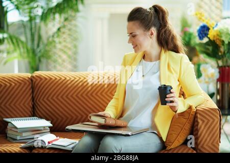 femme apprenant élégante en jean et veste jaune assise sur canapé avec un ordinateur portable fermé et café prêt à étudier en ligne dans la maison moderne en ensoleillé d Banque D'Images