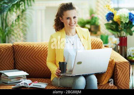 femme moderne souriante de 40 ans en jean et veste jaune dans le salon moderne en journée ensoleillée avec contenu pédagogique numérique sur un ordinateur portable. Banque D'Images