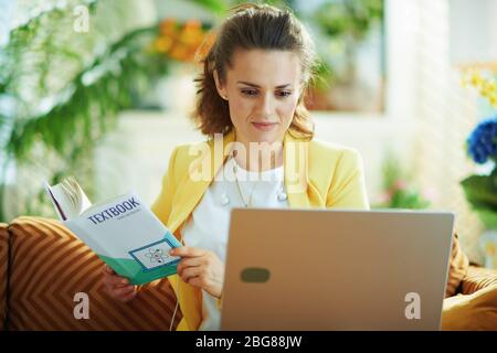 étudiant élégant en jean et veste jaune dans le salon moderne en journée ensoleillée en utilisant le site web d'e-learning sur un ordinateur portable. Banque D'Images