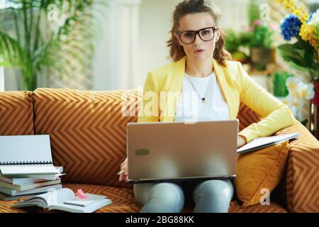 jeune femme apprenant en jean et veste jaune dans le salon moderne en journée ensoleillée regardant l'atelier numérique sur un ordinateur portable. Banque D'Images