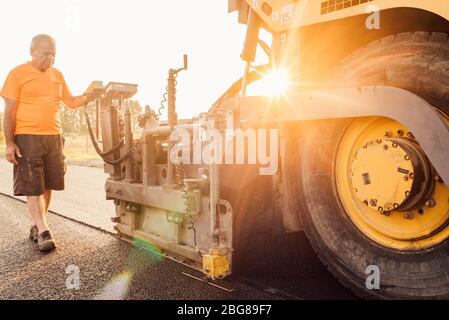 Travailleur routier exploitant une machine à asphalte qui pond de l'asphalte ou du bitume neuf pendant la construction de routes Banque D'Images