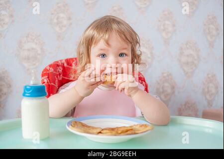 Petite fille mangeant des crêpes avec du lait dans un bavoir rose. Joli petit bébé mangeant des crêpes. Le bébé mange de manière indépendante. Assis dans une chaise haute. Banque D'Images