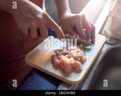 Gros plan à l'aide d'un couteau tranchant, trancher le poulet sur un panneau de découpe en bois près de la fenêtre dans la chambre en copropriété le soir. Cuisine à la maison. Banque D'Images