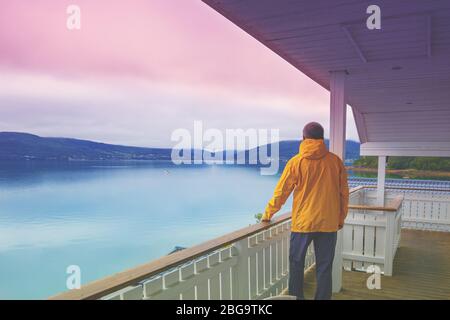 Le jeune homme debout sur la terrasse et regardant la mer. Vue panoramique sur le fjord. Le crépuscule avec le ciel rose. Beau paysagiste de montagne Banque D'Images