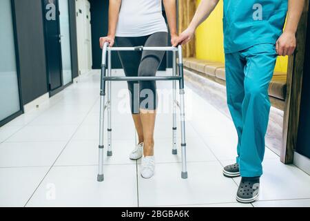 Femme qui utilise un marcheur, marche le long du couloir d'une clinique moderne de réadaptation. Le Réhabilitologue aide le patient à marcher. Réhabilita après blessure Banque D'Images