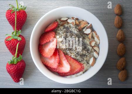 Flocons d'avoine végétalien sain avec fraises, graines de chia et amandes tranchées dans un bol blanc avec fraises et amandes entières en arrière-plan Banque D'Images