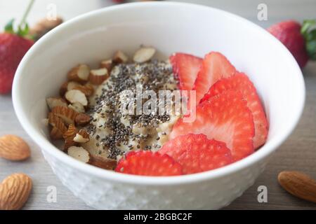 Flocons d'avoine végétalien sain avec fraises, graines de chia et amandes tranchées dans un bol blanc avec fraises et amandes entières en arrière-plan Banque D'Images