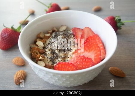 Flocons d'avoine végétalien sain avec fraises, graines de chia et amandes tranchées dans un bol blanc avec fraises et amandes entières en arrière-plan Banque D'Images