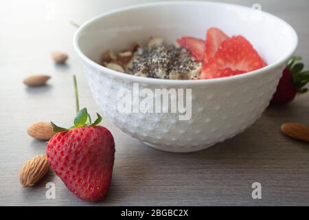 Flocons d'avoine végétalien sain avec fraises, graines de chia et amandes tranchées dans un bol blanc avec fraises et amandes entières en arrière-plan Banque D'Images