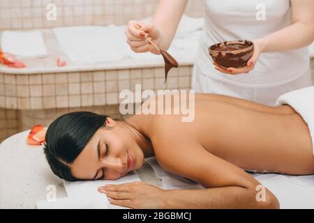 Femme mixte de course avec masque de corps en chocolat dans le salon de spa. Complexe de luxe et massage. Gommage au chocolat Banque D'Images