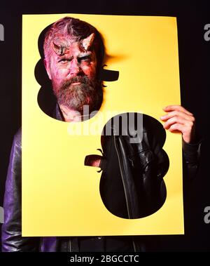 Concept de fête d'Halloween. Le diable ou monstre célèbre le festival. L'homme regarde la silhouette de citrouille coupée sur fond noir. Démon avec cornes et visage sérieux tient l'affiche jaune avec des cadres sculptés Banque D'Images