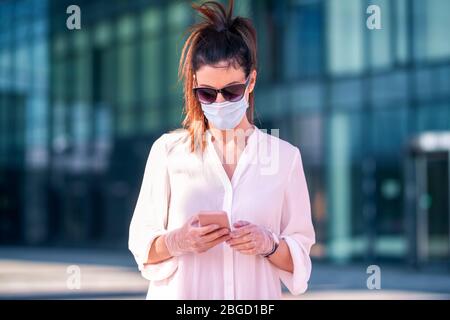 Gros plan sur une femme sûre d'elle portant un masque facial et des messages texte sur son téléphone mobile tout en marchant devant le centre d'affaires pendant le coronavirus Banque D'Images