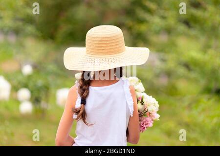 Une fille avec des piquets tient dans ses mains un bouquet de pivoines blanches et roses. Un enfant dans un chapeau de paille à large bord, le jour ensoleillé de l'été. Vert backgrou Banque D'Images