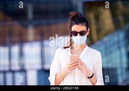 Gros plan sur une femme sûre d'elle portant un masque facial et des messages texte sur son téléphone mobile tout en marchant devant le centre d'affaires pendant le coronavirus Banque D'Images