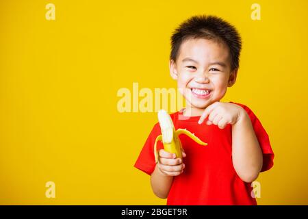 Joyeux portrait enfant ou enfant asiatique mignon petit garçon beau sourire portant un t-shirt rouge jouant tient pelé banane pour manger, studio tourné isolé sur Banque D'Images