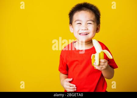 Joyeux portrait enfant ou enfant asiatique mignon petit garçon beau sourire portant un t-shirt rouge jouant tient pelé banane pour manger, studio tourné isolé sur Banque D'Images