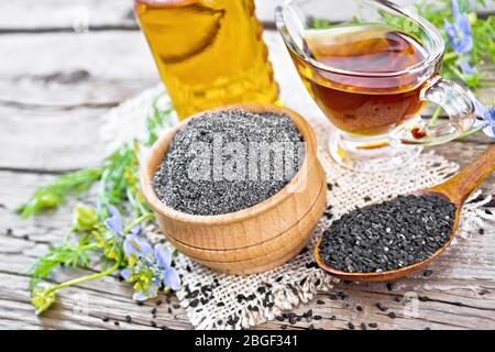 Farine de caraway noir dans un bol, graines dans une cuillère, huile en bouteille et sauceboat sur napkin de burlap, branches de Nigella sativa avec fleurs bleues et vert Banque D'Images