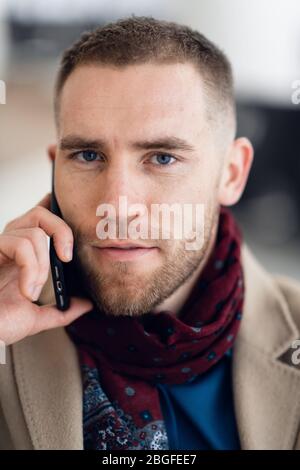 Jeune homme sérieux portant un foulard et un manteau parlant sur un téléphone portable gros plan. Banque D'Images