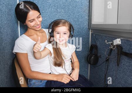 La petite fille est assise sur le circuit de sa mère pendant un test auditif, montre un pouce dans le bureau de l'audiologiste. Audiogramme, examen pour enfants dans l'oreille Banque D'Images