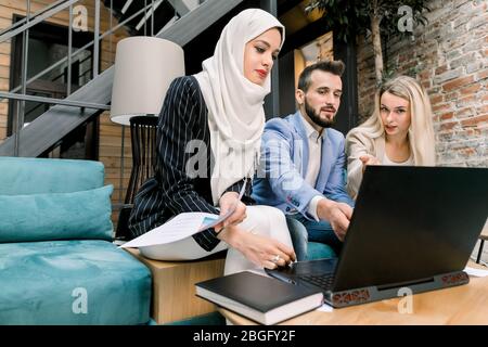 Trois jeunes collègues multiethniques, caucasiens, homme et femme, musulmane dans le hijab, ayant une conversation et utilisant un ordinateur portable, assis sur un canapé bleu Banque D'Images