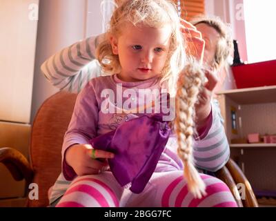 Une fille de deux ans (presque trois) est vérifiée pour les poux de tête par sa mère. Elle tient une poupée de Rainnzel Banque D'Images