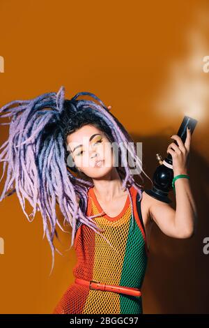 La femme rastafarienne fume de la marijuana. Bong fumeur de cannabis. Femme en robe rasta et rouge violet sur fond orange Banque D'Images