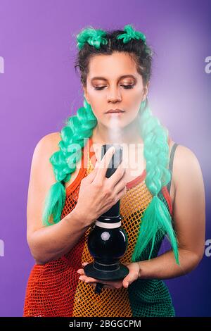 Grande bong en verre entre les mains d'une femme taille basse. La femme fume le cannabis. Coiffure féminine avec kanekalon vert sur fond violet Banque D'Images