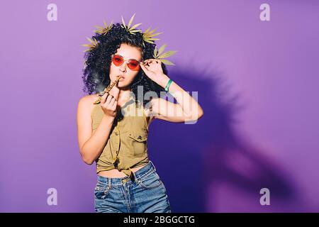 La femme Hipster fume la marijuana par un tuyau spécial pour fumeurs. Une femme contribue à la légalisation du cannabis. Portrait sur fond violet Banque D'Images