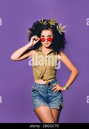 Femme mixte de course avec des feuilles de cannabis dans ses cheveux regarde le monde par des lunettes de soleil roses. Portrait sur fond violet. Légalisation de marijua Banque D'Images