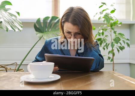 Femme mûre sérieuse prenant le petit déjeuner à la maison en pyjama avec une tasse de thé assis à la table de lecture tablette numérique. Triste femme de lire mail, nouvelles. NEG Banque D'Images