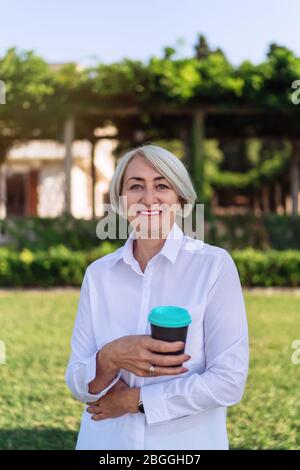 Heureuse femme mûre tenant le café à emporter tout en se reposant au parc Banque D'Images