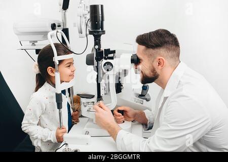 L'ophtalmologiste mâle vérifie la vue d'une petite fille avec un équipement ophtalmique spécial. GRL sourit parce que sa vue est très bonne Banque D'Images