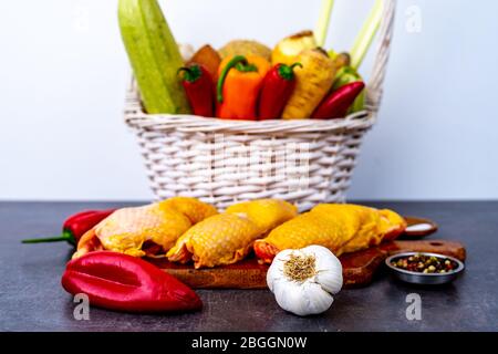 Panier plein de légumes et de racines derrière le tableau de découpe avec de la viande de poulet Banque D'Images