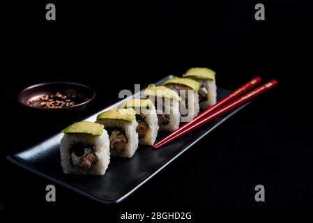 Rouleau de sushi avec wrap avocat, saumon grillé, fromage de Philadelphie et oignons verts sur plaque noire et baguettes rouges. Petit pot avec sauce soja. Noir Banque D'Images