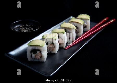 Rouleau de sushi avec wrap avocat, saumon grillé, fromage de Philadelphie et oignons verts sur plaque noire et baguettes rouges. Petit pot avec sauce soja. Noir Banque D'Images