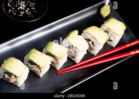 Rouleau de sushi avec wrap avocat, saumon grillé, fromage de Philadelphie et oignons verts sur plaque noire et baguettes rouges. Petit pot avec sauce soja. Noir Banque D'Images