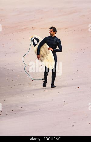 Surfeur masculin portant une combinaison, tenant planche de surf sous son bras, marchant sur la plage après la séance de surf du matin. Banque D'Images