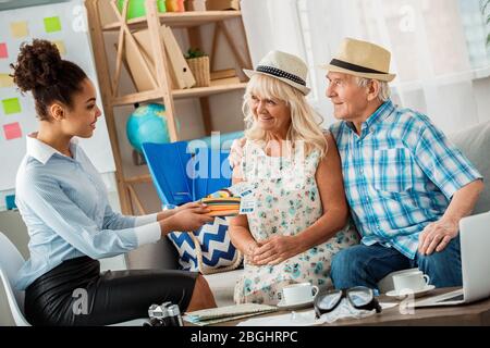 L'agent de voyage au bureau se réjouit de donner des passeports et des billets aux clients seniors dans des chapeaux de plage assis vue latérale Banque D'Images