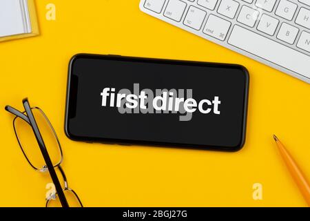Un smartphone affichant le logo First Direct repose sur un fond jaune avec un clavier, des lunettes, un stylo et un livre (usage éditorial uniquement). Banque D'Images